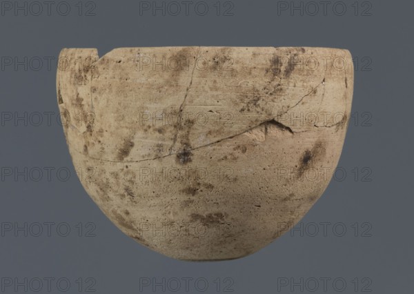 Bowl, unknown northern Mesopotamian workshop, Early Jazira III period ca. 2600–ca. 2350 BC Early Dynastic III period, clay vessel; bowl with an egg-shaped body, rounded bottom and a straight, non-separated rim slightly bent inwards; ca. 2600–ca. 2350 BC; Northern Mesopotamia Khabur basin, Tell Rad Shakra, North-Eastern Syria; vessel; height 7.8 cm, width 10.7 cm, A rounded, beige ceramic bowl with an uneven rim and visible cracks and chips sits against a solid gray background; the bowl surface shows darker brown smudges and spots, areas of lighter off-white patches, and fine horizontal striations in the clay texture.