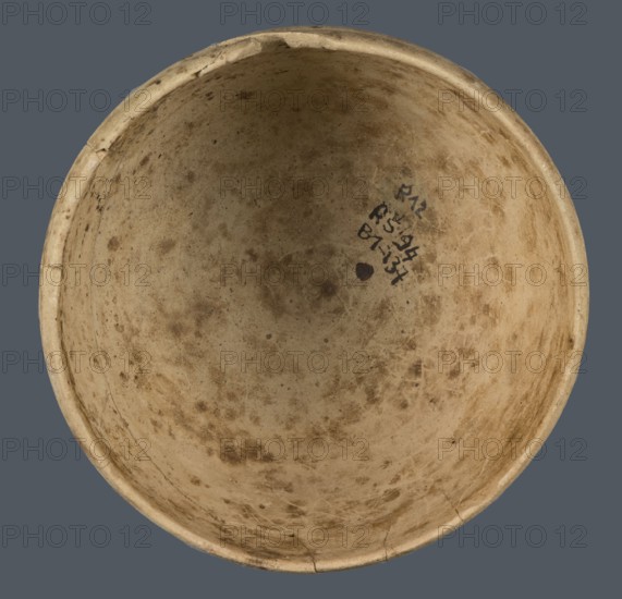 Bowl, unknown northern Mesopotamian workshop; Early Jazira III period ca. 2600–ca. 2350 BC / Early Dynastic III period; clay vessel; bowl with an egg-shaped body, rounded bottom, and a straight, non-separated rim slightly bent inwards; ca. 2600–ca. 2350 BC; Northern Mesopotamia Khabur basin; Tell Rad Shakra north-eastern Syria, Syria; vessel; height 7.8 cm; width 10.7 cm, A round, shallow bowl viewed from above showing an interior surface with a worn, mottled texture and small areas of discoloration; the rim is slightly uneven and has a few chips and hairline cracks; there is a small dark mark and a cluster of darker smudges near one side; the background is a solid muted gray. The visible colors include beige, tan, brown, dark brown, and gray.