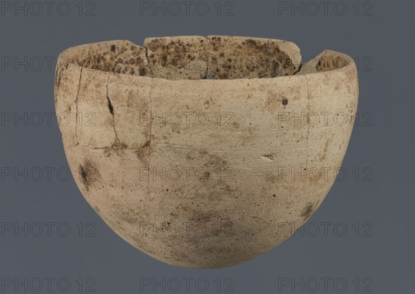 Bowl, unknown northern Mesopotamian workshop, Early Jazira III period ca. 2600–ca. 2350 BC Early Dynastic III period. Clay vessel; bowl with an egg-shaped body, rounded bottom, and a straight, non-separated rim slightly bent inwards. Northern Mesopotamia Khabur basin, Tell Rad Shakra North-Eastern Syria, Syria. ca. 2600–ca. 2350 BC. Height 7.8 cm; width 10.7 cm., A roughly hemispherical ceramic vessel with a worn, uneven rim that shows a few chips and cracks, displaying a weathered surface texture with small pits and scratches; the exterior is primarily light beige with areas of darker brown and tan discoloration and several small dark spots, while the interior rim has mottled brown staining; the background is a solid muted gray-blue.