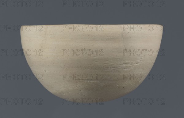 Bowl, unknown Northern Mesopotamian workshop; clay; Early Jazira III period ca. 2600–ca. 2350 BC / Early Dynastic III period. Tableware vessel with a flat, slightly oblique oval bottom with traces of being cut off with a string, gently tilted walls that straighten and bend slightly inwards in the upper part, and a straight, undifferentiated rim. Northern Mesopotamia Khabur basin, Tell Rad Shakra Northeastern Syria., A rounded bowl with a smooth, matte surface in shades of beige and light tan set against a solid dark gray background; the bowl shows subtle horizontal striations, fine surface speckles, and a few shallow scratches and hairline cracks, with slight variations in tone creating areas of warmer cream and cooler gray-beige.