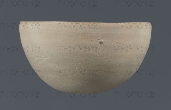Bowl, unknown Northern Mesopotamian workshop; clay; Early Jazira III period ca. 2600–ca. 2350 BC / Early Dynastic III period; tableware vessel from Northern Mesopotamia Khabur basin. Bowl with a flat, slightly oblique, oval bottom with traces of being cut off with a string, gently tilted walls that straighten out and bend slightly inwards in the upper part, and a straight, undistinguished rim. Tell Rad Shakra Northeastern Syria., A smooth, rounded bowl with a matte, pale beige surface showing subtle variations in tone and texture sits against a muted bluish-gray background; the bowl has a slightly uneven rim, areas of faint scratches and tooling marks, and small pitted imperfections, with visible shades of off-white, cream, light tan, and hints of gray.