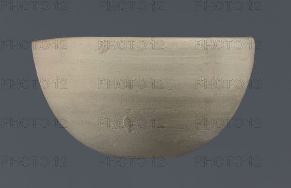 Bowl, unknown Northern Mesopotamian workshop, clay, Early Jazira III period ca. 2600–ca. 2350 BC / Early Dynastic III period. Tableware bowl with a flat, slightly oblique, oval bottom with traces of being cut off with a string, gently tilted walls which straighten out and bend slightly inwards in the upper part of the vessel, straight rim. Origin: Northern Mesopotamia Khabur basin, Tell Rad Shakra Northeastern Syria., A simple rounded vessel with a smooth matte surface fills the frame against a solid dark gray background, showing a pale creamy beige color with subtle variations into light tan and faint gray tones; the rim appears slightly darker with a hint of warm peach, and the body displays small darker speckles, fine horizontal striations, and a few shallow pits and scratches that create gentle surface texture.