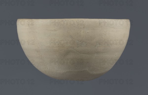 Bowl, unknown Northern Mesopotamian workshop, clay, Early Jazira III period ca. 2600–ca. 2350 BC Early Dynastic III period. Tableware bowl with a flat, slightly oblique, oval bottom with traces of being cut off with a string, gently tilted walls that straighten and bend slightly inwards in the upper part, and a straight, undistinguished rim. Origin: Al-Jazira Khabur basin, Northern Mesopotamia Tell Rad Shakra, northeastern Syria., A smooth rounded bowl with a matte surface suspended against a flat background, the bowl showing variations of beige and light tan with subtle darker speckles and faint irregular streaks, and the background appearing as a uniform muted bluish-gray.