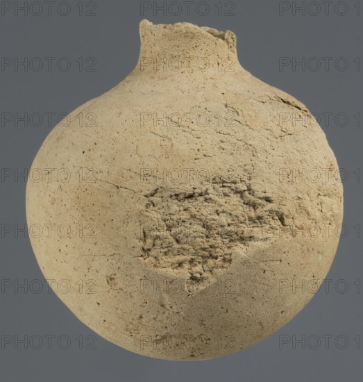 Globular vessel, unknown Northern Mesopotamian workshop; Early Jazira III period ca. 2600–ca. 2350 BC / Early Dynastic III period ca. 2600–ca. 2350 BC. Globular vessel with a narrow neck the upper part of the neck and the spout are not preserved. Thickened circular base. Clay; vessels for oils and ointments; vice-free vessel. Origin: Al-Jazira, Northern Mesopotamia Khabur basin, Tell Rad Shakra, Syria. Height 11.3 cm; width 10.45 cm., A round, weathered clay vessel with a narrow, broken neck sits against a muted gray background; the vessel surface is predominantly beige with patches of light tan and dusty brown, showing rough, porous texture, fine cracks, and an area of crumbly, uneven erosion with layered fragments, and subtle speckles and darker tiny spots scattered across the curved surface.