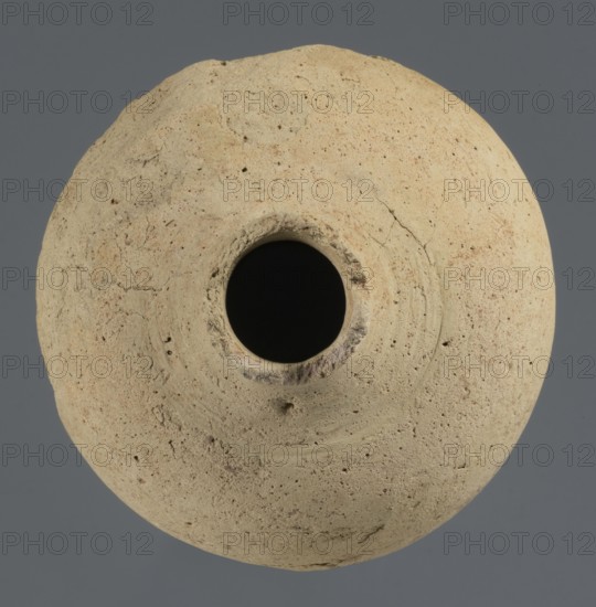 Globular vessel, unknown Northern Mesopotamian workshop. Early Jazira III period / Early Dynastic III period ca. 2600–ca. 2350 BC. Globular vessel with a narrow neck the upper part of the neck and the spout are not preserved. Thickened circular base. Clay vessel for oils and ointments; vice-free. Origin: Al-Jazira, Northern Mesopotamia Khabur basin, Tell Rad Shakra north-eastern Syria. Height 11.3 cm; width 10.45 cm., A round, weathered clay object with a central circular opening viewed from above, showing a rough, pitted surface in beige and light tan tones with small darker specks and subtle reddish-beige patches, set against a smooth gray background.