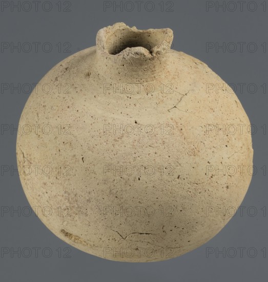 Globular vessel, unknown Northern Mesopotamian workshop, Early Jazira III period ca. 2600–ca. 2350 BC Early Dynastic III period. Globular vessel with a narrow neck the upper part of the neck and the spout are not preserved. Thickened circular base. Clay; vessels for oils and ointments; vice-free vessels. Origin: Al-Jazira Khabur basin, Northern Mesopotamia; Tell Rad Shakra, Syria. Height 11.3 cm; width 10.45 cm., A round, weathered ceramic vessel with a rough, pitted surface in pale beige and light tan tones sits against a muted gray background; the vessel has an irregular, flared opening with a scalloped rim showing darker beige shadows, and the body displays subtle reddish-brown speckling, small dark pits, and a few hairline cracks.