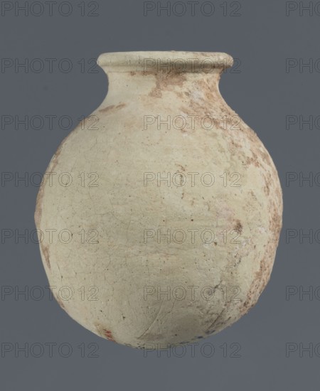 Spherical vessel, unknown northern Mesopotamian workshop, clay, Early Jazira III / Early Dynastic III period ca. 2600–ca. 2350 BC. Spherical vessel with an egg-shaped belly with a rounded bottom and a low cylindrical neck ending with a thickened rim turned outwards. Origin: Northern Mesopotamia Khabur basin, Tell Rad Shakra, Syria. Vessel; height 7.8 cm, width 6.5 cm., A rounded clay vessel with a narrow neck and flared rim sits against a solid gray background; the vessel surface is mostly off-white with areas of pale beige and light brown discoloration, visible weathering and surface cracks, and patches of darker brown and muted reddish-brown along one side, with a small area of faint blue and a touch of red near the lower edge.