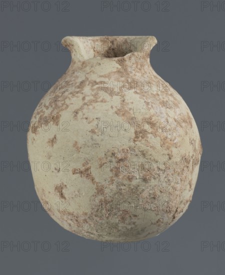 Spherical vessel, unknown northern Mesopotamian workshop; clay; Early Jazira III period ca. 2600–ca. 2350 BC / Early Dynastic III; spherical vessel with an egg-shaped belly with a rounded bottom and a low cylindrical neck ending with a thickened rim turned outwards; Northern Mesopotamia Khabur basin; Tell Rad Shakra North-Eastern Syria, Syria; height 7.8 cm, width 6.5 cm., A rounded, worn ceramic vessel with a narrow flared rim is centered against a solid background; the surface of the vessel shows an uneven, mottled texture with patches of light beige, pale cream, and muted brown, and areas of rough, flaky encrustation and worn, pitted spots are visible across its curved body.