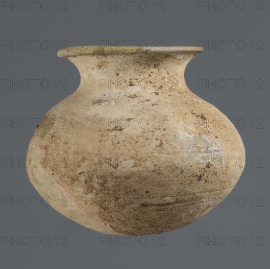 Vessel with an ellipsoidal belly, unknown northern Mesopotamian workshop, Early Jazira III period ca. 2600–ca. 2350 BC / Early Dynastic III period ca. 2600–ca. 2350 BC, clay vessel for oils and ointments, a small vessel with an ellipsoidal belly, rounded bottom, low flared neck ending in a flat top thickened by an outward curl; ca. 2600–ca. 2350 BC; Tell Rad Shakra, Northern Mesopotamia Khabur basin, Al-Jazira — Syria/Iraq, height 7.4 cm, width 8.6 cm., A rounded, bulbous vessel with a short neck and flared rim sits against a solid gray background; the vessel surface appears rough and weathered with a mottled palette of beige, tan, light brown, and patches of pale green and whitish discoloration, showing areas of wear, small pits, and uneven texture across its curved body.