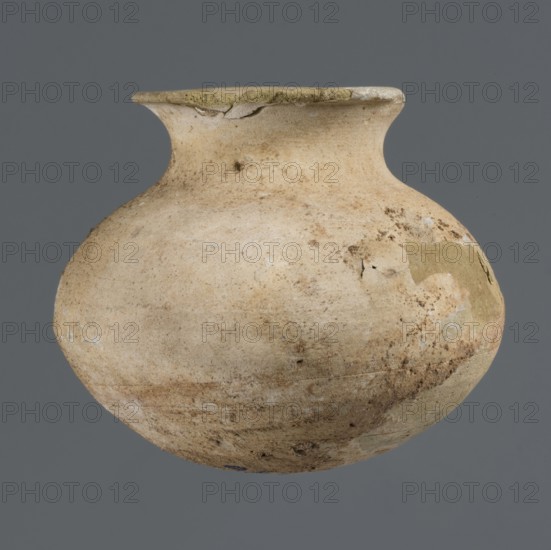 Vessel with an ellipsoidal belly, unknown northern Mesopotamian workshop, Early Jazira III period ca. 2600–ca. 2350 BC / Early Dynastic III period ca. 2600–ca. 2350 BC. Small clay vessel for oils and ointments with an ellipsoidal belly, rounded bottom, low flared neck ending in a flat top thickened by an outward curl. Origin: Northern Mesopotamia Al-Jazira, Khabur basin, Tell Rad Shakra, Syria. Height 7.4 cm; width 8.6 cm., A rounded ceramic vessel with a short neck and flared rim sits against a plain gray background; the surface shows a weathered, uneven texture with patches of lighter and darker beige, tan, and ochre, along with areas of brown discoloration and small spots of darker mottling, and a few rough or chipped edges near the rim.