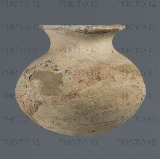 Vessel with an ellipsoidal belly, unknown northern Mesopotamian workshop; Early Jazira III / Early Dynastic III period ca. 2600–ca. 2350 BC; clay vessel for oils and ointments; small vessel with an ellipsoidal belly, rounded bottom, low flared neck ending in a flat top thickened by an outward curl; Al-Jazira northern Mesopotamia, Khabur basin, Tell Rad Shakra Northeastern Syria; height 7.4 cm, width 8.6 cm., A rounded ceramic vessel with a narrow neck and flared rim floats against a solid gray background, showing an uneven surface texture with patches of rough, worn areas. The vessel displays a palette of beige, tan, pale brown, and muted olive-green, with some areas of lighter off-white and faint reddish-brown discoloration. The surface shows irregular marks, scratches, and flaking, and a few cracked or chipped spots along the rim and body. The overall shape is bulbous with a constricted throat leading to the rim, and the finish appears matte and weathered.