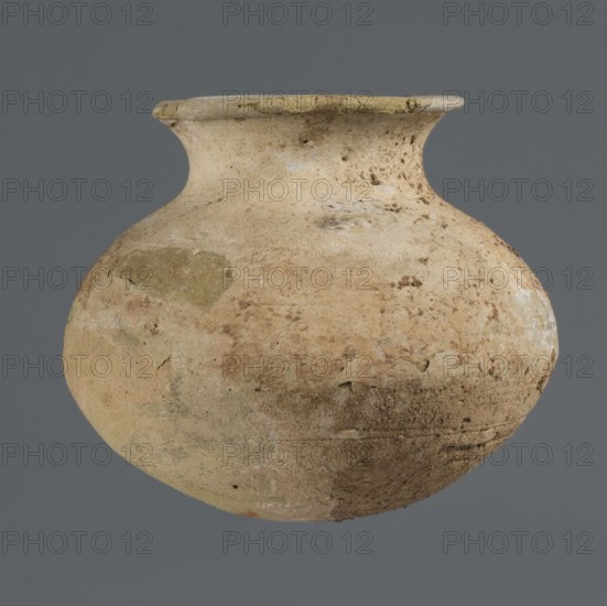 Vessel with an ellipsoidal belly, unknown northern Mesopotamian workshop, Early Jazira III period ca. 2600–ca. 2350 BC / Early Dynastic III period ca. 2600–ca. 2350 BC; clay vessel for oils and ointments; a small vessel with an ellipsoidal belly, rounded bottom, low flared neck ending in a flat top, thickened by an outward curl; Al-Jazira Khabur basin, Northern Mesopotamia Tell Rad Shakra, northeastern Syria; height 7.4 cm, width 8.6 cm., A weathered rounded clay vessel with a narrow neck and flared rim sits against a flat gray background; the pot displays a rough, pitted surface with patches of lighter beige, tan, pale yellow, and muted green, along with areas of brown discoloration and faint whitish deposits, and its texture shows cracks, chips, and uneven wear.