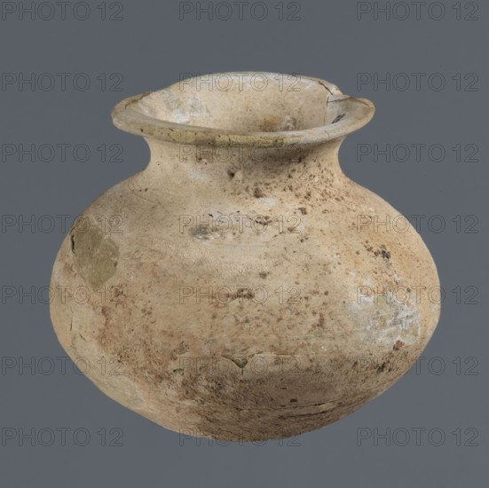 Vessel with an ellipsoidal belly, unknown northern Mesopotamian workshop, Early Jazira III period ca. 2600–ca. 2350 BC / Early Dynastic III period ca. 2600–ca. 2350 BC, clay vessel for oils and ointments, small vessel with an ellipsoidal belly, rounded bottom, low flared neck ending in a flat top thickened by an outward curl, provenance: Tell Rad Shakra, Khabur basin, Northeastern Syria Northern Mesopotamia, height 7.4 cm, width 8.6 cm., A rounded clay vessel with a flared rim shown against a solid gray background, the vessel surface bearing an overall beige to light tan color with areas of darker brown spotting and patches of pale off-white and greenish discoloration, the rim appearing slightly lighter with small chips and a crack, the body showing rough texture, worn and uneven surfaces, and a few small dark specks and smudges.