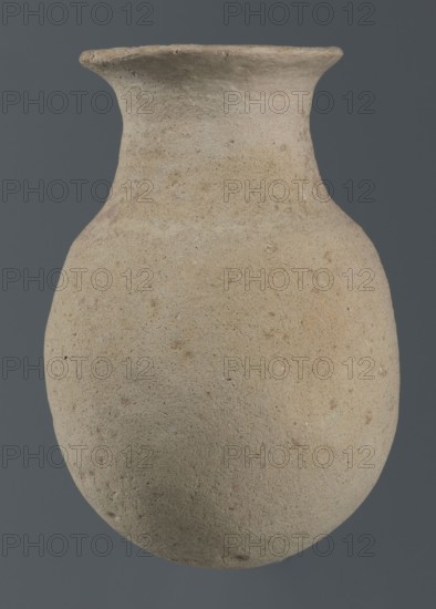 Flask, unknown northern Mesopotamian workshop, Early Jazira IV period ca. 2350–ca. 2200 BC / Akkadian period ca. 2350–ca. 2200 BC. Flask-shaped vessel with an egg-shaped belly, a rounded bottom, a high cylindrical neck separated from the belly by narrow shoulders and ending with a flat, tilted rim. ca. 2350–ca. 2200 BC. Northern Mesopotamia Khabur basin; Tell Rad Shakra, Syria. Vessel; height 14.5 cm, width 9 cm., A single rounded vessel with a narrow neck and flaring rim fills the frame, showing a coarse, textured surface with small pits and irregularities; the vessel appears in shades of beige, tan, light brown, and hints of pale gray, set against a solid dark gray background.