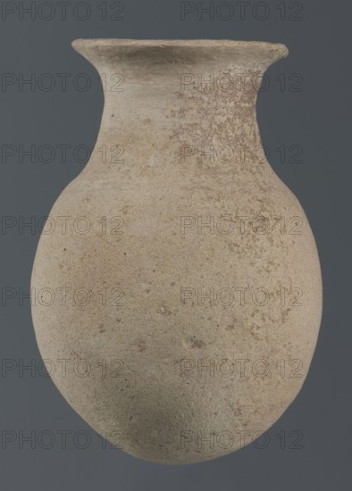 Flask, unknown northern Mesopotamian workshop; vice-free vessels, tableware; Early Jazira IV period ca. 2350–ca. 2200 BC / Akkadian period ca. 2350–ca. 2200 BC. Flask-shaped vessel with an egg-shaped belly, a rounded bottom, a high cylindrical neck separated from the belly by narrow shoulders and ending with a flat, tilted rim. ca. 2350–ca. 2200 BC. Syria, Northern Mesopotamia Khabur basin; Tell Rad Shakra Northeastern Syria. Vessel; height 14.5 cm, width 9 cm., A rounded, bulbous clay vessel with a narrow neck and flared rim sits against a solid dark gray background; the surface of the vessel shows a rough, grainy texture with a mix of beige, light tan, and pale brown tones and areas of slightly darker brown spotting and mottling.
