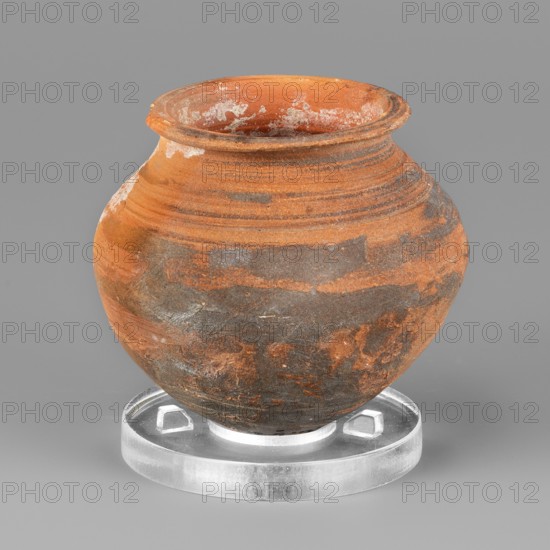 Vessel, unknown northern Mesopotamian workshop, metallic ware, viceless vessels, clay vessels, vessels for oils and ointments, miniature vessel with an ovoid belly with a pointed bottom and a low neck terminating in a narrow, upturned rim. 1st half of the 3rd millennium BC; Early Jazira III period, Northern Mesopotamia Khabur Basin, Tell Rad Shakra Northeastern Syria. Vessel; height 5.5 cm, width 6.1 cm, weight 60 g., An orange-brown rounded clay vessel with a flared rim sits on a clear circular stand, showing areas of darker grayish-brown discoloration and patches of whitish residue on its rim and body, with horizontal striations around the upper portion and an overall textured, slightly weathered surface against a smooth gray background.