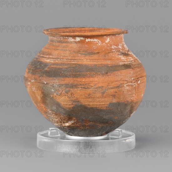 Vessel, unknown northern Mesopotamian workshop, metallic ware; viceless vessel for oils and ointments; clay miniature vessel with an ovoid belly, pointed bottom, low neck terminating in a narrow upturned rim. 1st half of the 3rd millennium BC; Early Jazira III period, Northern Mesopotamia Khabur Basin, Tell Rad Shakra Northeastern Syria. Height 5.5 cm, width 6.1 cm, weight 60 g., A round ceramic pot with a flared rim sits on a clear circular stand; the pot shows a textured surface with a mix of orange and dark gray tones forming layered, streaky patterns, plus small areas of white residue and lighter beige patches, all set against a neutral gray background.