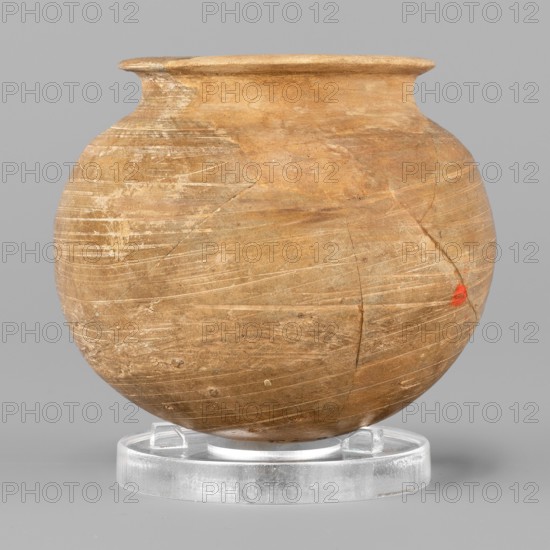 Vessel, unknown Northern Mesopotamian workshop; low vessel with a spherical belly, rounded bottom and low, wide neck with a narrow, upturned spout. Smoothed and fired in a reduction atmosphere. 1st half of the 3rd millennium BC; Early Jazira III period, Northern Mesopotamia Khabur basin, Tell Rad Shakra North-eastern Syria. Function: vessels for oils and ointments; spherical vessel. Material/culture: vice-free vessels; material culture. Height 6.1 cm, width 6.9 cm, weight 85 g., A round, wide-mouthed pot with a bulbous body sits on a clear circular stand; the pot's surface shows a weathered, earthy tan and brown coloration with visible lighter scratch-like marks and a small area of bright red on one side, and the stand and background present shades of clear/translucent and light gray respectively.