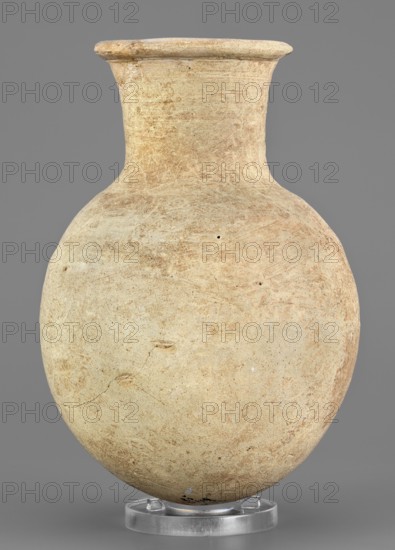 Flask, unknown Northern Mesopotamian workshop. Clay flask-shaped vessel with an elongated ovoid belly, rounded bottom, high slightly conically flaring neck separated from the belly by narrow shoulders and ending with a flat top, protruding externally thickened rim with a triangular cross-section. 2nd half of the 3rd millennium BC; Early Jazira IV Akkadian period, ca. 2350–2200 BC. Northern Mesopotamia Khabur basin, Tell Rad Shakra North-Eastern Syria. Height 14.8 cm, width 10.2 cm, weight 315 g., A rounded vessel with a narrow neck and flared rim sits upright on a clear circular stand; the surface of the vessel shows a textured, worn appearance with patches of lighter and darker beige and tan, along with subtle reddish-brown areas and small darker specks; the background is a flat medium gray.