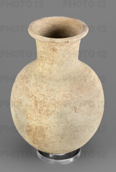 Flask, unknown Northern Mesopotamian workshop. Clay flask-shaped vessel with an elongated ovoid belly, rounded bottom, high slightly conically flaring neck separated from the belly by narrow shoulders, ending with a flat top and a protruding externally thickened rim with a triangular cross-section. 2nd half of the 3rd millennium BC; Early Jazira IV period Akkadian period; ca. 2350–ca. 2200 BC. Northern Mesopotamia Khabur basin, Tell Rad Shakra North-Eastern Syria. Height 14.8 cm, width 10.2 cm, weight 315 g., A rounded ceramic vessel with a narrow neck flaring into a wide rim sits against a plain gray background; the vessel shows a textured surface with uneven patches of light beige, tan, and pale brown, with some slightly darker brown areas and faint surface marks and speckles, and it rests on a clear circular stand that appears colorless and slightly reflective.