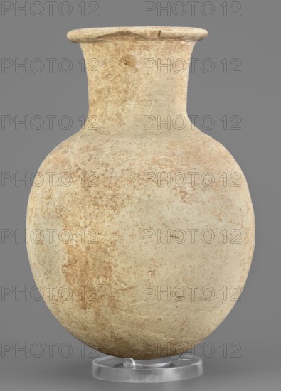 Flask, unknown Northern Mesopotamian workshop; clay flask-shaped vessel with an elongated ovoid belly, rounded bottom, high slightly conically flaring neck separated from the belly by narrow shoulders and ending with a flat top; protruding externally thickened rim with triangular cross-section; 2nd half of the 3rd millennium BC, Early Jazira IV Akkadian period, ca. 2350–ca. 2200 BC; Northern Mesopotamia Khabur basin, Tell Rad Shakra North-Eastern Syria; height 14.8 cm, width 10.2 cm, weight 315 g., The image shows a rounded ceramic vessel with a narrow neck and a slightly flared rim, displayed upright on a clear circular stand against a plain gray background; the vessel surface is textured and worn with a mix of beige, tan, light brown, and pale cream tones, and there are areas of darker brown discoloration and subtle surface blemishes.