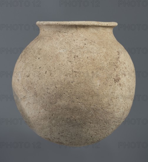 Spherical vessel, unknown Northern Mesopotamian workshop, Early Jazira III period ca. 2600–ca. 2350 BC, clay; vice-less spherical vessel with a rounded bottom. In the upper part the vessel wall bends inward, creating sloping arms that merge into an arched, low neck terminating in a narrow, outwardly obliquely cut rim with a triangular cross section. c. 2600–ca. 2350 BC Early Dynastic III period. Northern Mesopotamia Khabur basin, Tell Rad Shakra, Northeastern Syria. Vessel; height 20.3 cm, width 21 cm., A rounded clay vessel with a narrow neck and outward-turned rim fills the frame, showing a textured surface with irregular pitting and small flecks; the vessel appears in shades of beige, tan, and light brown with subtle darker brown speckles, set against a uniform medium gray background.