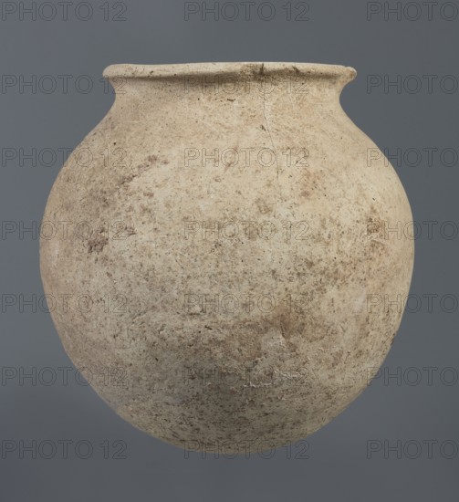 Spherical vessel, unknown Northern Mesopotamian workshop, Early Jazira III period ca. 2600–ca. 2350 BC Early Dynastic III period, clay, vice-less spherical vessel with a rounded bottom. In the upper part of the vessel wall the vessel bends inwards, creating sloping arms that merge into an arched, low neck terminating in a narrow, outwardly obliquely cut rim with a triangular cross-section. c. 2600–ca. 2350 BC; Northern Mesopotamia Khabur basin, Tell Rad Shakra Northeastern Syria. Height 20.3 cm; width 21 cm., A large rounded clay pot with a narrow rim sits against a plain gray background; the pot's surface is textured and rough with a mix of beige, tan, brown, and off-white tones, showing patches of darker brown and subtle speckles, and a visible hairline crack near the rim.