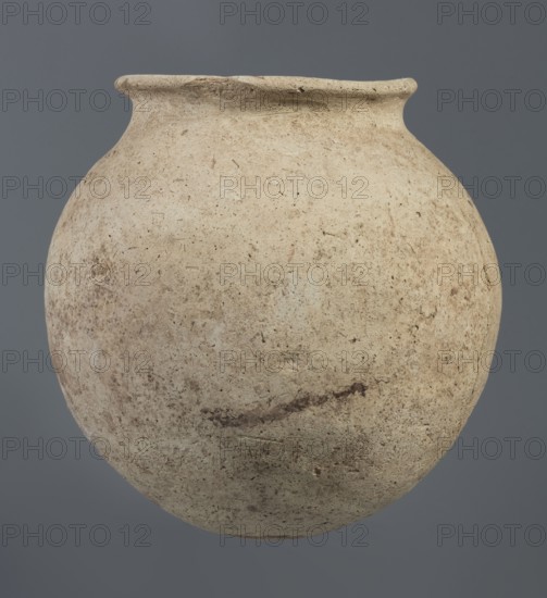 Spherical clay vessel, vice-less, from an unknown Northern Mesopotamian workshop, Early Jazira III / Early Dynastic III period ca. 2600–ca. 2350 BC. Vice-less spherical vessel with rounded bottom; upper wall bends inward forming sloping arms that merge into an arched low neck terminating in a narrow, outwardly obliquely cut rim with a triangular cross-section. Origin: Northern Mesopotamia Al-Jazira, Khabur basin, Tell Rad Shakra, Northeastern Syria. Height 20.3 cm, width 21 cm., A rounded, weathered clay vessel with a narrow flared rim fills the frame against a solid gray background, showing a rough, pitted surface with a palette of beige, tan, cream, light brown, and hints of darker brown; the vessel displays irregular speckling, smudges, and subtle linear marks across its body and rim, with variations in texture from smoother upper areas to more grainy, mottled lower sections.