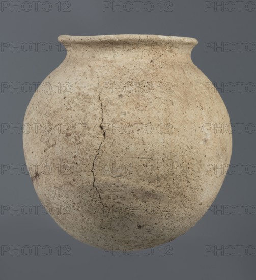 Spherical vessel, unknown Northern Mesopotamian workshop, Early Jazira III period ca. 2600–ca. 2350 BC Early Dynastic III period. Clay, vice-less spherical vessel with rounded bottom. In the upper part the vessel wall bends inward, creating sloping arms that merge into an arched, low neck terminating in a narrow, outwardly obliquely cut, triangular-cross-section rim. c. 2600–ca. 2350 BC; Northern Mesopotamia Khabur basin, Al-Jazira, Tell Rad Shakra, northeastern Syria. Height 20.3 cm; width 21 cm., A large rounded vessel with a narrow flared rim occupies the frame, showing a textured, weathered surface with visible cracks and small pits; the vessel is mostly beige and light tan with darker brown speckles and areas of discoloration, set against a uniform gray background.
