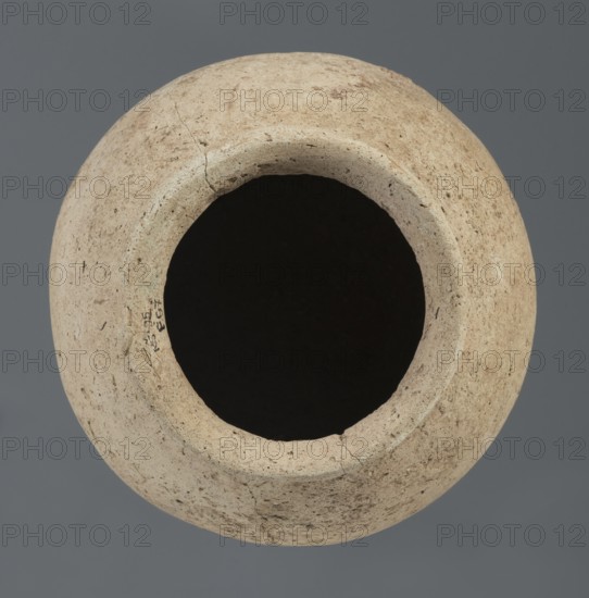 Spherical vessel, unknown Northern Mesopotamian workshop; Early Jazira III period ca. 2600–ca. 2350 BC; Early Dynastic III period; vice-less spherical vessel with rounded bottom. In the upper part the vessel wall bends inward, creating sloping arms that merge into an arched, low neck terminating in a narrow, outwardly obliquely cut rim with a triangular cross-section. Material: clay. Origin: Northern Mesopotamia Khabur basin, Tell Rad Shakra, Northeastern Syria. Height 20.3 cm; width 21 cm., A round, weathered clay vessel seen from above with a wide circular opening revealing a dark interior, the outer surface showing a rough, pitted texture with cracks and small imperfections, the vessel displaying shades of beige, tan, cream, and light brown against a muted gray background.