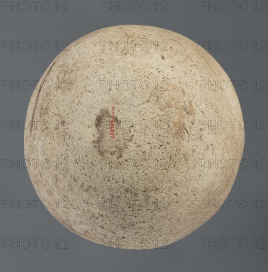 Spherical vessel, unknown Northern Mesopotamian workshop; Early Jazira III period ca. 2600–ca. 2350 BC; Early Dynastic III period. Clay vessel, vice-less spherical vessel with a rounded bottom. The upper part of the vessel wall bends inward, creating sloping arms that merge into an arched, low neck terminating in a narrow, outwardly obliquely cut rim with a triangular cross-section. c. 2600–ca. 2350 BC. Northern Mesopotamia Khabur basin, Tell Rad Shakra Northeastern Syria. Height 20.3 cm; width 21 cm., A large round object fills the frame against a muted gray background, showing a rough, pitted surface in shades of beige, cream, and light tan with patches of darker brown and subtle gray speckling; there are a few irregular darker smudges and a thin crack near the top, and the overall texture appears granular and weathered.