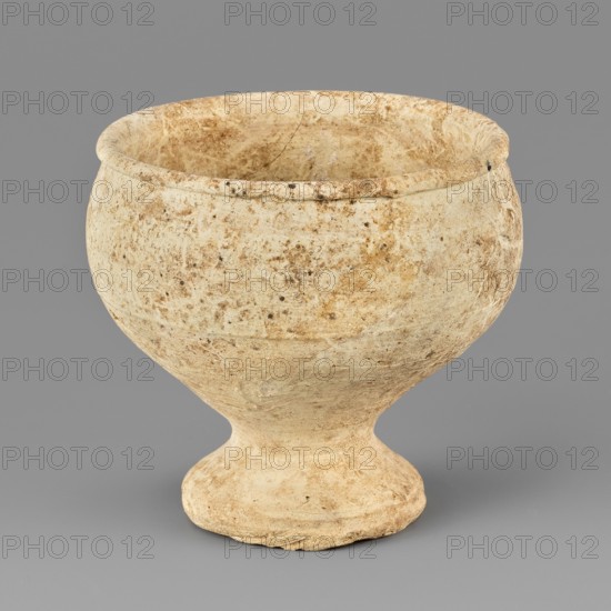 Footed bowl, unknown Northern Mesopotamian workshop. Miniature bowl on a low, conically formed circular foot, flat on the underside. The bowl walls are conical, straightening out at two-thirds of their height, bending gently towards the interior; they terminate in a low, rounded rim that turns outward. Clay vessel, footed bowl. 1st half of the 3rd millennium BC; Early Jazira III period Early Dynastic III; ca. 2600–ca. 2350 BC. Tell Rad Shakra North-Eastern Syria, Northern Mesopotamia Khabur basin. Height 4.8 cm, width 5.3 cm., A rounded, bowl-shaped vessel with a flared rim and a pedestal base sits against a plain background; the surface shows a mottled, weathered texture with tones of beige, tan, light brown, and hints of off-white, and there are a few darker speckles and irregular patches across the exterior.