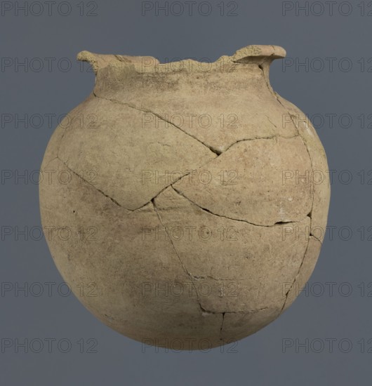Spherical vessel, unknown Northern Mesopotamian workshop, Early Jazira III period ca. 2600–ca. 2350 BC. Spherical vessel with an egg-shaped belly and a rounded bottom; in the upper part the belly merges into a low cylindrical neck terminating in a narrow, strongly protruding, rounded rim turned outwards. Vice-free bulbous clay vessel. Early Dynastic III period ca. 2600–ca. 2350 BC. Al-Jazira Khabur basin, Northern Mesopotamia; Tell Rad Shakra northeastern Syria, c. 2600–ca. 2350 BC. Height 16.5 cm, width 16.2 cm., A rounded, earthen-colored ceramic vessel with a textured surface and visible repaired cracks occupies the image against a solid gray background; the vessel shows a rim with two small outward flaring projections and uneven, chipped edges, and its surface displays variations of beige, tan, light brown, and pale yellow tones with darker brown areas along some cracks and edges.