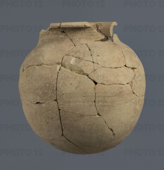 Spherical vessel, unknown Northern Mesopotamian workshop, Early Jazira III period ca. 2600–ca. 2350 BC. Spherical vessel with an egg-shaped belly and a rounded bottom; in the upper part the belly merges into a low cylindrical neck terminating in a narrow, strongly protruding, rounded rim turned outwards. Clay vessel; vice-free, bulbous type. Origin: Northern Mesopotamia Khabur basin, Tell Rad Shakra northeastern Syria. Height 16.5 cm; width 16.2 cm., A rounded ceramic vessel shown against a solid grey background, composed of many joined clay fragments with visible cracks and seams; the vessel has a bulbous body and a broken, irregular rim with a small raised lip, and its surface shows a rough, matte texture. The colors visible include various shades of beige, tan, light brown, and hints of pale ochre, with subtle darker brown speckling and slightly lighter areas along some edges and seams.