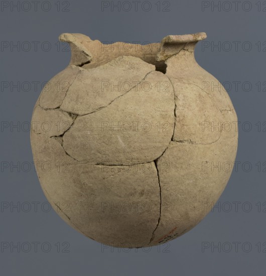 Spherical vessel, unknown Northern Mesopotamian workshop, Early Jazira III period ca. 2600–ca. 2350 BC / Early Dynastic III period ca. 2600–ca. 2350 BC. Spherical vessel with an egg-shaped belly and a rounded bottom; in the upper part the belly merges into a low cylindrical neck terminating in a narrow, strongly protruding, rounded rim turned outwards. Clay bulbous, vice-free vessel. Origin: Al-Jazira Khabur basin, Northern Mesopotamia; Tell Rad Shakra northeastern Syria. c. 2600–ca. 2350 BC. Height 16.5 cm; width 16.2 cm., A rounded, light beige clay vessel with a rough, matte surface and visible repaired cracks fills the frame against a flat, muted gray background, showing a flared, uneven rim with a few chipped edges and a series of irregular, slightly darker fracture lines and patched joins across its body, with subtle variations of tan, sandy brown, and faint grayish tones on the pot's surface.