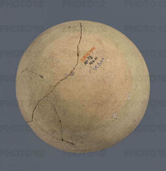 Spherical vessel, unknown Northern Mesopotamian workshop, Early Jazira III period ca. 2600–ca. 2350 BC / Early Dynastic III period ca. 2600–ca. 2350 BC. Spherical vessel with an egg-shaped belly and a rounded bottom; in the upper part the belly merges into a low cylindrical neck terminating in a narrow, strongly protruding, rounded rim turned outwards. Clay, vice-free bulbous vessel. Origin: Al-Jazira Khabur basin, Northern Mesopotamia; Tell Rad Shakra northeastern Syria. Height 16.5 cm, width 16.2 cm., A round, weathered object with a rough, sandy surface in shades of beige and tan, showing a few dark cracks and hairline fractures across its surface; there is a small area with traces of darker markings in muted blue and red, and a few tiny pale spots and scuffs; the background is a solid muted gray.