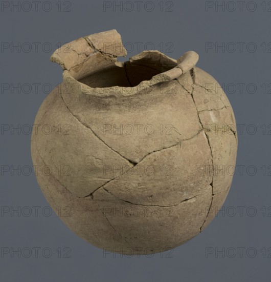 Spherical vessel, unknown Northern Mesopotamian workshop; Early Jazira III period ca. 2600–ca. 2350 BC; Early Dynastic III period ca. 2600–ca. 2350 BC. Spherical vessel with an egg-shaped belly and a rounded bottom. In the upper part the belly merges into a low cylindrical neck terminating in a narrow, strongly protruding, rounded rim turned outwards. Clay vessel; vice-free, bulbous form. Origin: Northern Mesopotamia Khabur basin, Tell Rad Shakra, northeastern Syria. c. 2600–ca. 2350 BC. Height 16.5 cm; width 16.2 cm., A weathered, round pottery vessel with a broken rim and missing fragments showing visible cracks and repaired joins, displaying a rough, uneven surface texture; the vessel is primarily tan and beige with areas of darker brown and traces of lighter off-white, set against a plain gray-blue background.
