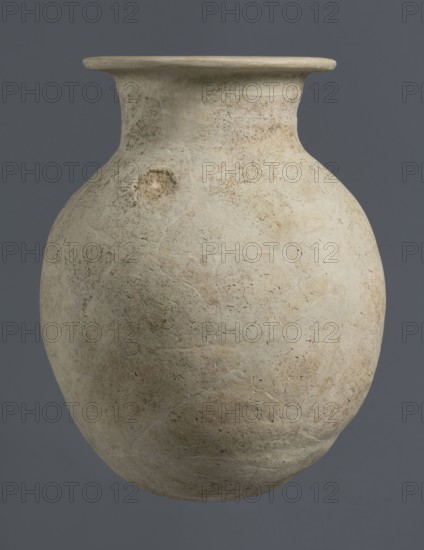 Vise-less jug, unknown northern Mesopotamian workshop. Early Jazira III / Early Dynastic III period, ca. 2600–2350 BC. Clay vessel: vise-less jug with an ovoid belly, flat crookedly cut round bottom, sloping shoulders rising to a high cylindrical neck terminating in a wide flat spout protruding outwards. Bulbous vessel; jug. Origin: Northern Mesopotamia Khabur basin, Al-Jazira. Found at Tell Rad Shakra northeastern Syria., A large rounded clay vessel with a wide flared rim sits against a solid dark gray background. The vessel's surface is a pale beige to off-white with areas of light brown and faint tan discoloration, showing a textured, slightly pitted and weathered finish. There are visible repaired seams and subtle irregularities in shape, including a rough circular patch on one side. The rim is smoother and lighter in tone than the body, and the overall appearance is matte with small speckles and faint mottling of beige, tan, and light brown.