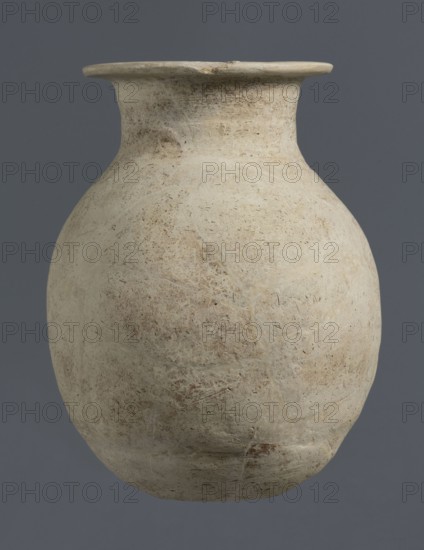 Vise-less jug, unknown northern Mesopotamian workshop; Early Jazira III period ca. 2600–ca. 2350 BC; clay; bulbous ovoid belly with a flat, crookedly cut round bottom; sloping shoulders leading to a high cylindrical neck terminating in a wide, flat outward-protruding spout; vise-less vessel; jugs; Al-Jazira Syria/Iraq, Mesopotamia; Early Dynastic III period ca. 2600–ca. 2350 BC; found at Tell Rad Shakra northeastern Syria, Northern Mesopotamia Khabur basin., A large rounded ceramic vessel with a narrow neck and flared rim sits against a solid dark gray background; the vessel shows a weathered, matte surface in shades of off-white, beige, and light tan with visible rough texture, small pits, and mottled darker brown areas, and faint irregular linear marks and patches suggesting surface wear.