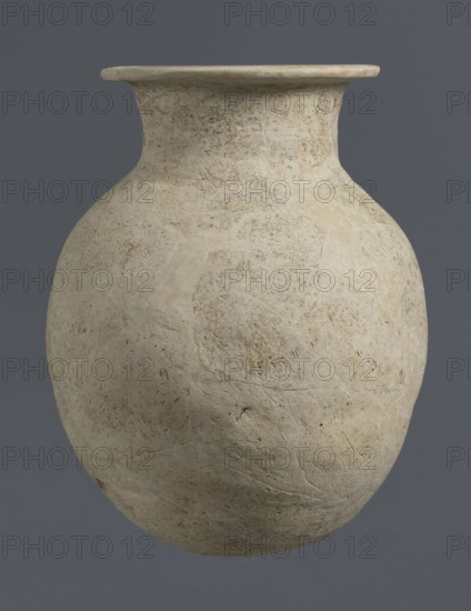 Vise-less jug, unknown northern Mesopotamian workshop; Early Jazira III period ca. 2600–ca. 2350 BC; clay; bulbous vessel — jug vise-less. Vise-less jug with an ovoid belly with a flat, crookedly cut round bottom, sloping shoulders, from which arises a high cylindrical neck terminating in a wide flat spout protruding outwards. Origin: Al-Jazira northern Mesopotamia, Khabur basin; discovered at Tell Rad Shakra, northeastern Syria., A large rounded vessel with a narrow neck and flared rim sits against a solid background; the vessel surface appears rough and weathered with an uneven texture and small pitting and scratches, showing a pale beige to off-white color with areas of light tan and faint gray mottling, while the background is a muted dark gray-blue.