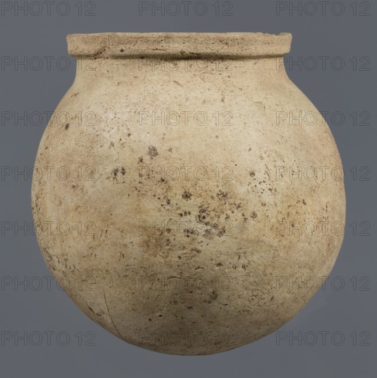 Spherical vessel, unknown northern Mesopotamian workshop, Early Jazira III period ca. 2600–ca. 2350 BC. A spherical vessel with a pear-shaped, tapering body with a rounded bottom, strongly sloping shoulders passing into a low, wide neck ending with a thickened rim on the outside in the form of a wide rectangular strip; the rounded outer edge protrudes above the inner surface of the neck, creating a lowered step for a lid. On the lower part of the body a pottery mark was carved before firing: two long lines intersecting at the bottom at an acute angle, forming a mark resembling a deformed letter Y. Material: clay; viseless pottery. Origin: Northern Mesopotamia Khabur basin, Tell Rad Shakra northeastern Syria. Date: c. 2600–c. 2350 BC Early Jazira III/Early Dynastic III. Dimensions: height 25.5 cm; width 24.6 cm., A large rounded clay vessel with a narrow flared rim fills the frame against a plain gray background; the vessel surface is an uneven beige to light tan with patches of darker ...