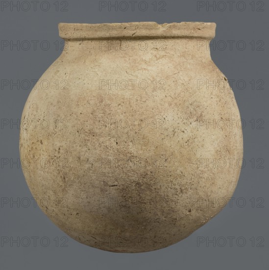 Spherical vessel, unknown northern Mesopotamian workshop. Early Jazira III period ca. 2600–ca. 2350 BC / Early Dynastic III period. A spherical vessel with a pear-shaped, tapering body with a rounded bottom, strongly sloping shoulders passing into a low, wide neck ending with a thickened rim on the outside in the form of a wide rectangular strip; the rounded outer edge protrudes above the inner surface of the neck, creating a lowered step for a lid. On the lower part of the body a pottery mark was carved before firing: two long lines intersecting at the bottom at an acute angle, forming a mark resembling a deformed letter Y. Origin: Northern Mesopotamia Khabur basin, Tell Rad Shakra northeastern Syria. Material: clay. Function: vessel. Height 25.5 cm; width 24.6 cm., A rounded clay vessel with a narrow rim displayed against a solid background, showing a textured surface with weathered marks and small pits; the vessel's coloration includes beige, tan, light brown, and hints of darker br...
