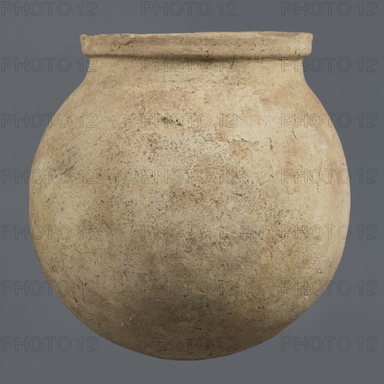 Spherical vessel, unknown northern Mesopotamian workshop, Early Jazira III period ca. 2600–ca. 2350 BC / Early Dynastic III period. A spherical pear-shaped, tapering body with rounded bottom, strongly sloping shoulders passing into a low, wide neck ending with a thickened rim formed as a wide rectangular strip whose rounded outer edge protrudes above the inner surface of the neck, creating a lowered step for a lid. On the lower part of the body a pottery mark was incised before firing: two long lines intersecting at the bottom at an acute angle, forming a deformed Y-shaped mark. Terracotta/clay vessel from Al-Jazira Khabur basin, Tell Rad Shakra northeastern Syria. Height 25.5 cm; width 24.6 cm., A rounded, bulbous ceramic vessel with a short, slightly flared neck and a thick rim sits against a flat background. The surface of the vessel is textured and worn, showing patches of roughness and small pits. The predominant color of the vessel is a light beige or tan with areas of darker bro...