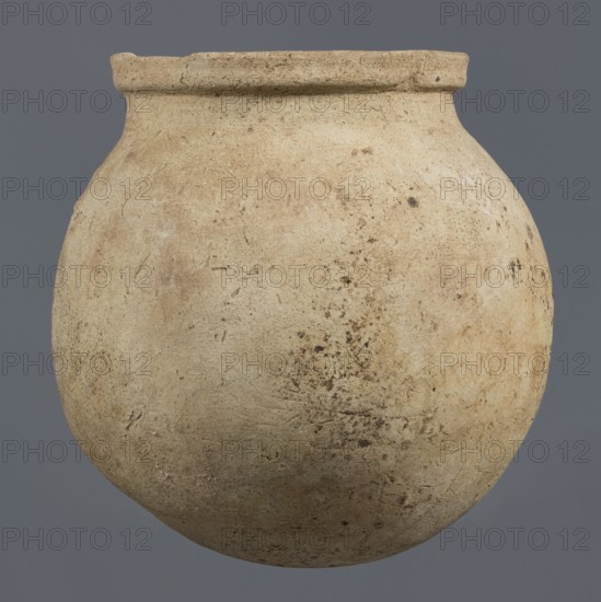 Spherical vessel, unknown northern Mesopotamian workshop, Early Jazira III period c. 2600–c. 2350 BC. A spherical vessel with a pear-shaped, tapering body with a rounded bottom, strongly sloping shoulders passing into a low, wide neck ending with a thickened rim on the outside in the form of a wide rectangular strip whose rounded outer edge protrudes above the inner surface of the neck, creating a lowered step for a lid. On the lower part of the body a pottery mark was carved before firing: two long lines intersecting at the bottom at an acute angle, forming a mark resembling a deformed letter Y. Materials: clay; type: viseless/ceramic vessel. Findspot: Tell Rad Shakra, Khabur basin, northern Mesopotamia Al-Jazira, Syria. Height 25.5 cm; width 24.6 cm., A rounded, bulbous ceramic vessel with a short, slightly flared neck and a narrow rim, shown against a flat background. The surface of the vessel is matte and textured with visible pits, scratches, and areas of uneven wear. The dominant...