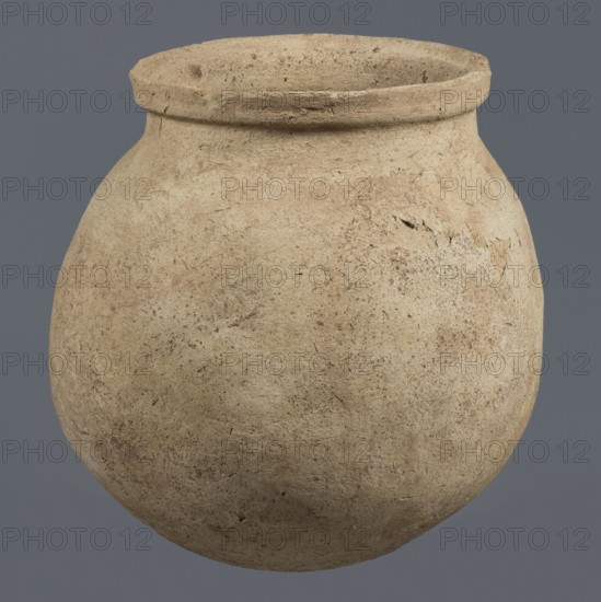 Spherical vessel, unknown northern Mesopotamian workshop, Early Jazira III period ca. 2600–ca. 2350 BC / Early Dynastic III period. A spherical vessel with a pear-shaped, tapering body with a rounded bottom, strongly sloping shoulders passing into a low, wide neck ending with a thickened rim on the outside in the form of a wide rectangular strip; the rounded outer edge of the rim protrudes above the inner surface of the neck, creating a lowered step for a lid. On the lower part of the body a pottery mark was carved before firing: two long lines intersecting at the bottom at an acute angle, forming a mark resembling a deformed letter Y. Material: clay. Origin: Northern Mesopotamia Al-Jazira/Khabur basin, Tell Rad Shakra northeastern Syria. Vessel; height 25.5 cm, width 24.6 cm., A large rounded clay vessel with a wide mouth and a thick rim, showing a rough, textured surface with small pits and irregularities, colored in shades of beige and light brown with darker brown speckles and patc...