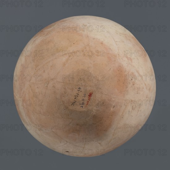 Spherical vessel, unknown northern Mesopotamian workshop; Early Jazira III / Early Dynastic III period ca. 2600–ca. 2350 BC. Clay, vice-free spherical vessel with an ovoid belly widening toward the top, small flat round bottom, short sloping shoulders merging into a low wide neck terminating in a rounded rim formed as a wide, thickened strip on the outside. Northern Mesopotamia Khabur basin, Tell Rad Shakra northeastern Mesopotamia, Syria. Height 28 cm; width 28 cm., A round, worn object with a smooth surface displaying shades of beige, tan, light brown, and pale peach sits against a solid gray background; the surface shows a few faint cracks, scuffs, and subtle variations in tone and texture, with a small darker circular area near the center and several tiny dark specks scattered across the surface.