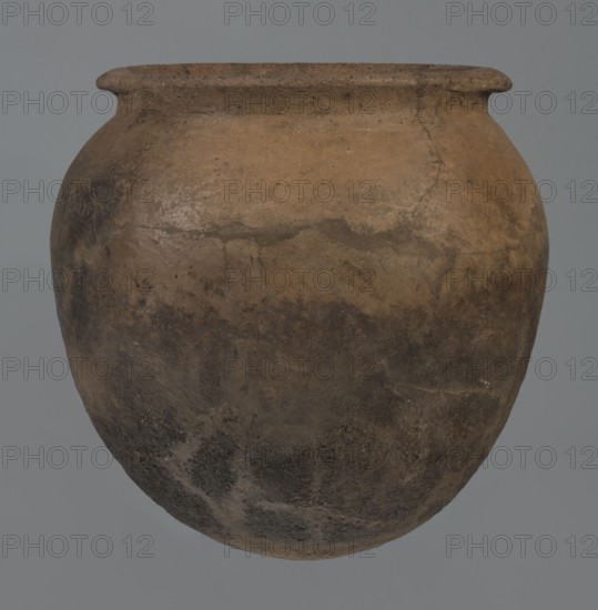 Kitchen pot, unknown northern Mesopotamian workshop, Early Jazira III period ca. 2600–ca. 2350 BC. Kitchen vessel with an egg-shaped belly and rounded bottom. Upper walls bend slightly inward to form narrow shoulders rising to a low cylindrical wide neck with a straight, slightly tilted spout. Two slightly thickened horizontal crescent-shaped handles attach on opposite edges. Outer walls intentionally roughened on the lower half and carefully polished above. Traces of soot visible on the bottom and slightly above. Syria; Tell Rad Shakra northeastern Syria. Vessel; height 20.5 cm, width 20.7 cm., A large rounded clay pot with a wide slightly flared rim shown against a plain gray background; the pot's surface displays an uneven, weathered texture with areas of light brown, tan, dark brown, and grayish-black, and visible mottled patches and subtle cracks across its body.