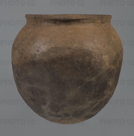 Kitchen pot, unknown northern Mesopotamian workshop, Early Jazira III period ca. 2600–ca. 2350 BC. A kitchen pot with an egg-shaped belly and rounded bottom. Upper walls bend slightly inward forming narrow shoulders that give rise to a low, cylindrical wide neck with a straight, slightly tilted spout. Two slightly thickened horizontal crescent-shaped handles are attached on opposite edges of the spout. Outer walls are intentionally roughened at the bottom up to half the vessel’s height and carefully polished above. Traces of soot are visible on the bottom and slightly above. Syria, Tell Rad Shakra northeastern Syria. Vessel; height 20.5 cm, width 20.7 cm., A large rounded clay pot with a narrow rim in shades of brown and tan, showing darker brown and almost black areas on its lower half and lighter tan patches near the top, with an overall textured, weathered surface and a muted gray background.