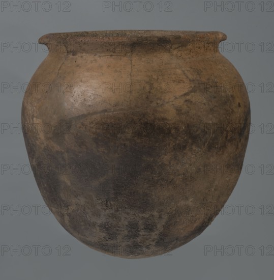 Kitchen pot, unknown northern Mesopotamian workshop; Early Jazira III period ca. 2600–ca. 2350 BC. A kitchen pot with an egg-shaped belly and a rounded bottom. In the upper part the walls bend slightly inwards, forming narrow shoulders that give rise to a low, cylindrical, wide neck with a straight, slightly tilted spout. Two slightly thickened horizontal crescent-shaped handles are attached on opposite edges. Outer walls are intentionally roughened at the bottom up to half the vessel’s height, while above they are carefully polished. Traces of soot are visible on the bottom and slightly above. Found at Tell Rad Shakra northeastern Syria. Vessel; height 20.5 cm, width 20.7 cm., A rounded, bulbous earthenware vessel with a slightly flared rim, showing an uneven surface with patches of brown, tan, dark brown, and muted black; the pot has a weathered, rough texture with visible discoloration and darker smudges concentrated toward the lower half, and the background is a flat gray.