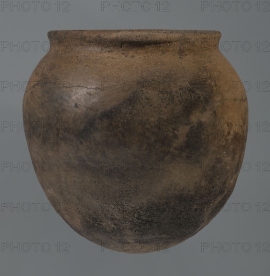 Kitchen pot, unknown northern Mesopotamian workshop, Early Jazira III period ca. 2600–ca. 2350 BC. A kitchen pot with an egg-shaped belly and a rounded bottom. In the upper part the walls bend slightly inward, forming narrow shoulders from which rises a low, cylindrical, wide neck with a straight, slightly tilted spout. Two slightly thickened horizontal crescent-shaped handles are attached on opposite edges. Outer walls are intentionally roughened at the bottom up to half the vessel’s height, while above they are carefully polished. Traces of soot are visible on the bottom and slightly above. Material: clay. Function: kitchen vessel/urn. Findspot: Tell Rad Shakra northeastern Syria. Dimensions: height 20.5 cm, width 20.7 cm., An old rounded clay vessel with a wide mouth and a slightly flared rim, showing an uneven, weathered surface in shades of brown, dark brown, tan, and gray, with mottled dark patches and lighter worn areas, subtle surface cracks and rough texture, set against a mut...